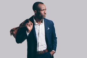 Confident in his perfect style. Handsome young African man in full suit carrying brown leather bag on shoulder and looking away while standing against grey background photo