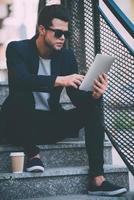 Surfing web outdoors. Handsome young man in smart casual wear working on digital tablet while sitting on stairs outdoors photo