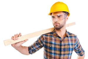 Confident contractor. Confident young male carpenter in hardhat and protective eyewear carrying wooden balk on shoulder while standing against white background photo