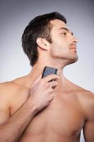 Shaving with electric shaver. Cropped image of handsome young man shaving his face with electric shaver while standing isolated on grey background photo