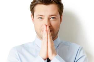 Praying for business. Portrait of thoughtful young man in shirt and tie holding hands clasped and looking at camera while standing isolated on white photo