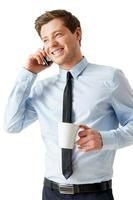 Time for coffee break. Happy young man in shirt and tie talking on the mobile phone and holding a cup of coffee while standing isolated on white photo