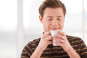 Having a coffee break. Handsome young man smelling coffee and keeping eyes closed while holding a cup near face photo