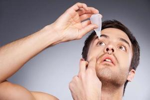 ojo goteando con gotas para los ojos. joven aplicando gotas para los ojos mientras está de pie contra el fondo gris foto