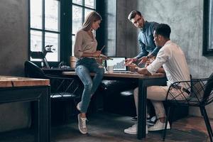 Three young people in smart casual wear analyzing data on laptop while working in office photo