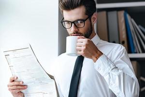 cinco minutos para café y periódico fresco. apuesto joven hombre de negocios con anteojos leyendo el periódico y bebiendo café mientras está en el cargo foto