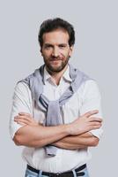 Confidence and style. Handsome young man keeping arms crossed and looking at camera while standing against grey background photo