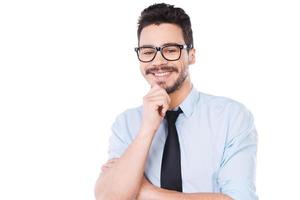 joven y exitoso. apuesto joven con camisa y corbata sosteniendo la mano en la barbilla y sonriendo mientras está de pie contra el fondo blanco foto