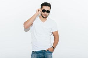 Confident and handsome. Handsome young Indian man adjusting his sunglasses and looking at camera while standing against white background photo