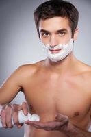 Man applying shaving cream. Handsome young shirtless man applying shaving cream at his face and looking at camera while standing isolated on grey background photo