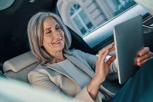 hermosa mujer madura con ropa informal inteligente usando una tableta digital mientras se sienta en el asiento trasero del auto foto