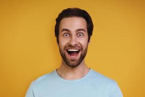 Portrait of excited young man in casual clothing looking at camera and smiling while standing against yellow background photo