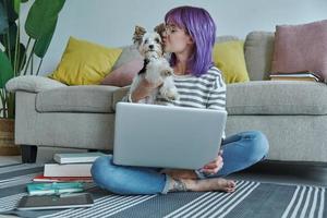 hermosa mujer joven con el pelo morado que lleva un lindo perrito mientras usa una laptop en casa foto