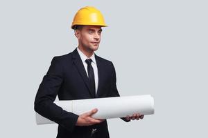 Young architect. Good looking young man in hardhat carrying blueprint and looking away while standing against grey background photo