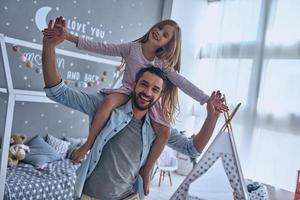 diversión sin preocupaciones. padre alegre cargando a su hija sobre los hombros y sonriendo mientras pasa tiempo libre en casa foto