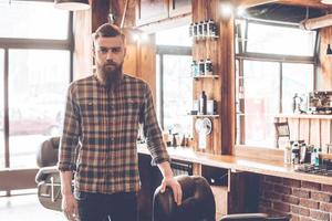 Confident barber expert. Young bearded man looking at camera and keeping hand on chair while standing at barbershop photo