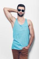 Confident and handsome. Confident young handsome man in sunglasses holding hand behind head and looking at camera while standing against white background photo