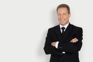 Confident businessman. Cheerful mature man in formalwear keeping arms crossed and smiling while standing against grey background photo