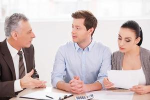 Financial consultation. Young couple sitting together at the table while senior man in formalwear telling something and gesturing photo