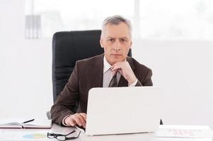 hombre de negocios confiado. un anciano seguro de sí mismo con ropa formal trabajando en una laptop y mirando la cámara mientras se sienta en su lugar de trabajo foto