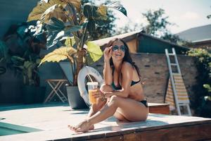 Attractive young woman in bikini drinking cocktail and smiling while sitting by the pool outdoors photo