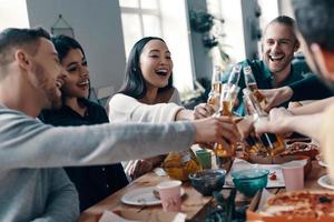 Friendship that lasts forever. Group of young people in casual wear toasting each other and smiling while having a dinner party indoors photo