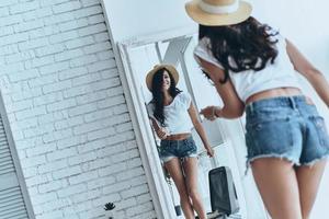 Ready for summer adventures. Rear view of attractive young woman trying on her sun hat and smiling while looking in the mirror at home photo