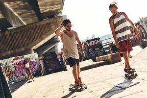 disfrutando de un día soleado. Longitud total de dos apuestos jóvenes patinando mientras pasan el rato con sus amigos al aire libre foto