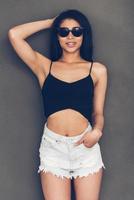 Beauty in style. Beautiful young woman in sunglasses holding hand behind head and looking at camera while standing against grey background photo