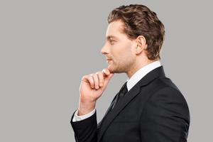 Generating new ideas. Side view of thoughtful young man in formalwear holding hand on chin and standing against grey background photo