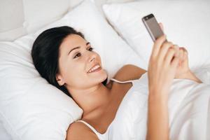 Always available for him. Beautiful young woman smiling and holding smart phone while lying in the bed at home photo