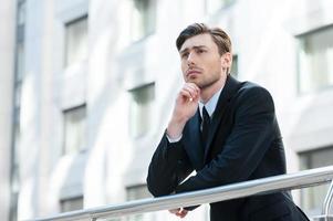 Thinking about solutions. Side view of young man in formalwear holding hand on chin and looking away while standing outdoors photo