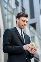 Young and wealthy. Confident young man in shirt and tie counting money and while standing outdoors photo