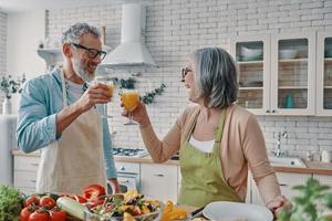 amorosa pareja de ancianos en delantales brindando con jugo de naranja y preparando una cena saludable mientras pasan tiempo en casa foto