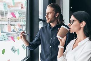 Passionate about their project. Two young colleagues in smart casual wear working while man using adhesive notes on the window photo