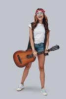 Full length of attractive stylish young woman playing acoustic guitar and smiling while standing against grey background photo