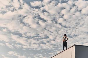 Her body earned a break. Modern young woman in sports clothing relaxing while standing on the roof outdoors photo