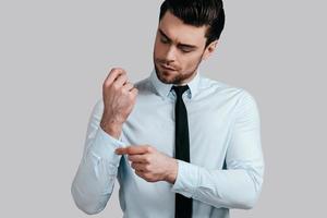 Always perfect. Handsome young man in white shirt and tie adjusting his sleeve while standing against grey background photo