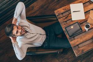 Enjoying his working day. Top view of handsome young man keeping hands behind head and looking at camera with smile while sitting at his working place photo