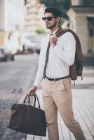 Handsome and stylish. Handsome young man in sunglasses holding leather bag and looking away while walking outdoors photo