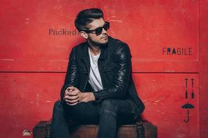 Gorgeous and stylish. Handsome young man in sunglasses keeping hands clasped and looking away while sitting on wooden chest against red background photo