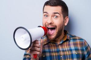 Want to be heard. Handsome young man shouting through a megaphone while standing against grey background. photo