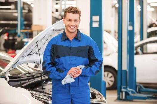 After the work is done. Confident young man in uniform wiping his hands with rag and smiling while standing in workshop with car in the background photo