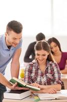 Studying in friendly atmosphere. Two cheerful students discussing something while looking at the book together with other students sitting behind them photo