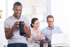 equipo creativo en el trabajo. apuesto joven africano sosteniendo la cámara y sonriendo mientras dos personas trabajan en segundo plano foto