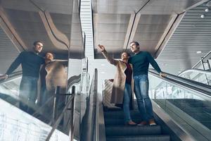 Mature couple pointing away and smiling while moving by escalator in airport terminal photo