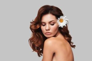 Pure beauty. Attractive young woman keeping chamomile in hair while standing against grey background photo