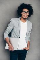 Keep on smiling. Cheerful young African man holding laptop and looking at camera with smile while standing against grey background photo