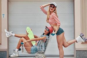 Having crazy day together. Side view of happy young woman on roller skates carrying her female friend in shopping cart and smiling while skating against the garage door photo
