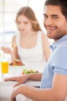 Starting morning from healthy breakfast. Handsome young man looking over shoulder and smiling while having breakfast with his girlfriend photo